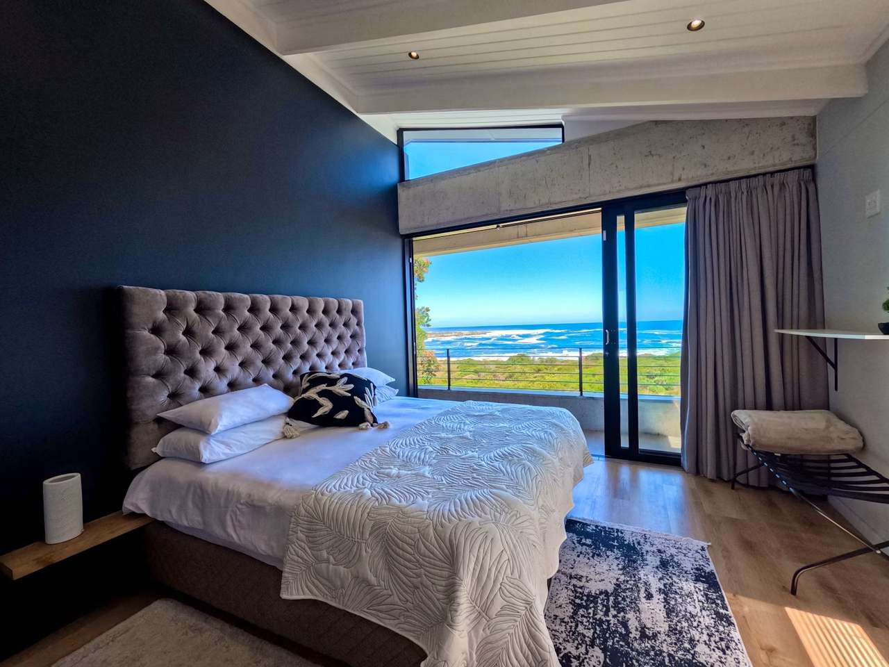 Upstairs ocean-view bedroom at Imoyo Ocean Villa with king bed, tufted headboard and balcony overlooking Walker Bay
