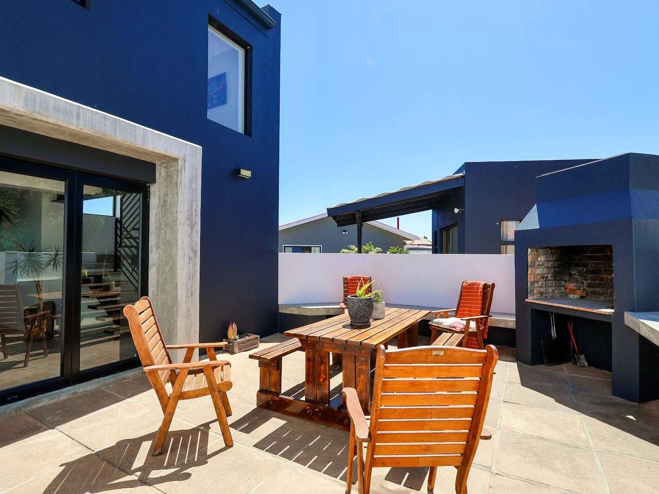 Sunny outdoor patio at Imoyo Ocean Villa with wooden dining table, chairs and built-in braai under blue sky